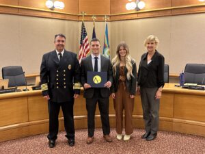 Ryan Gustin stands with Chief Andy Lay and Mayor Mary Ferdon during a swearing in ceremony at Columbus City Hall. Gustin holds folder containing his oath of office for the city. Standing beside Ryan is his wife. All are smiling for the photo. 